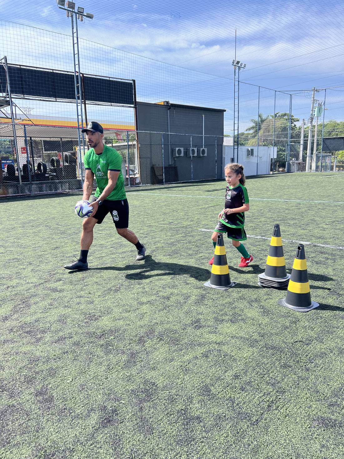 Professor da Arena Fut7 ao lado de um aluno, com bolas, cones e squeezes personalizados da escola
